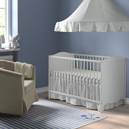 Una cuna SMÅGÖRA blanca con bordes biselados en una guardería azul, junto a un sillón gris y bajo un dosel blanco, con una alfombra de rayas azules.