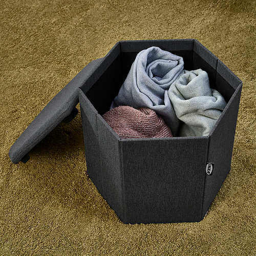 Una otomana de almacenamiento de tela gris con tapa extraíble sostiene la ropa doblada en un piso alfombrado.