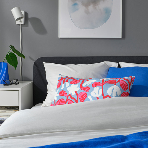 Cama moderna con almohada floral azul y roja, sábanas blancas. Planta y libro en mesa auxiliar con lámpara.
