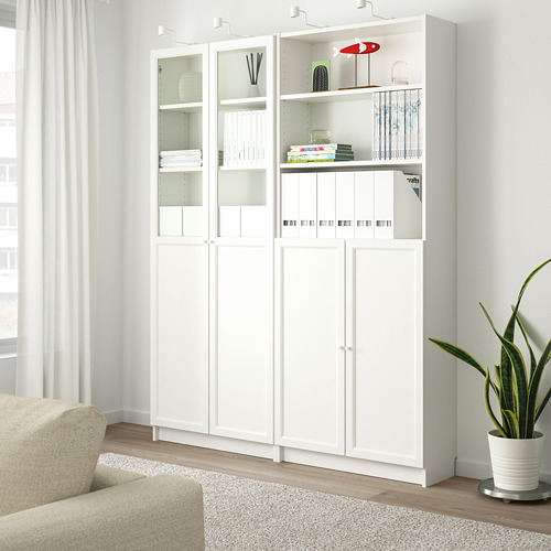 Librería blanca BILLY con puertas de cristal, con libros y decoración, junto a una silla beige y una gran maceta de macetas.