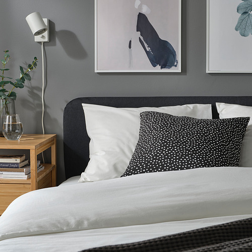 Acogedora cama gris con cojines blancos y punteados, arte de pared, planta pequeña, mesita de noche de madera con libros.