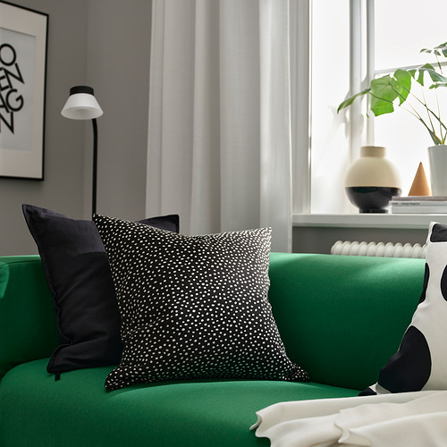 Elegante cojín verde BJÖRKGRÅMAL con estampado de puntos sobre una cama. Emparejado con cojín negro, contra pared gris con lámpara y planta. Decoración moderna.