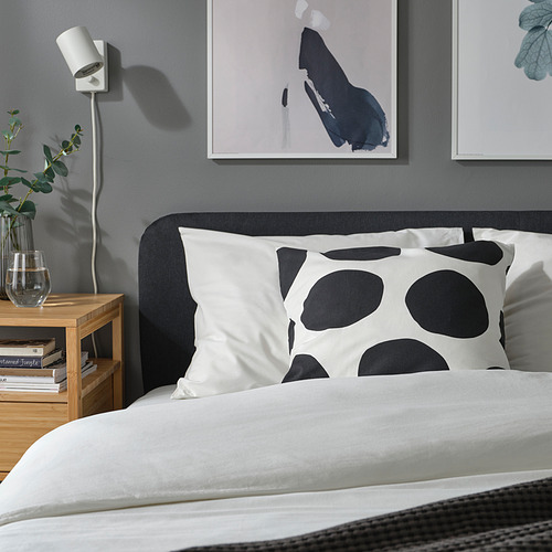 Dormitorio con almohada blanca con lunares negros, cabecero gris, decoración minimalista, arte abstracto y mesita de noche con jarrón y libros.