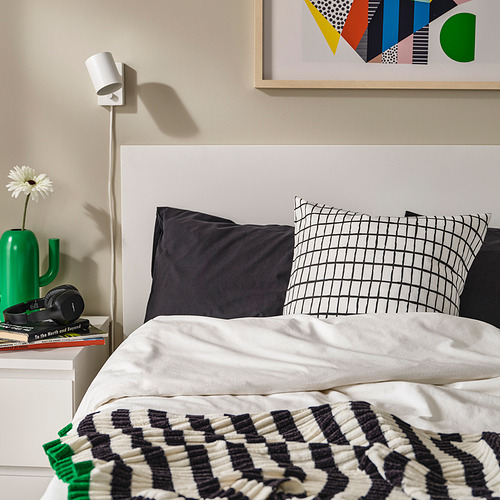 Cama con almohada a cuadros blancos y negros, jarrón verde, lámpara blanca, auriculares, libros, arte abstracto en la pared.