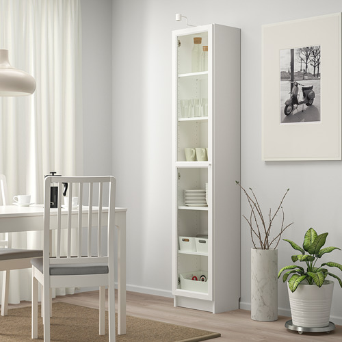 Estantería blanca BILLY con puertas de cristal, mostrando platos y vasos junto a una silla y un jarrón.