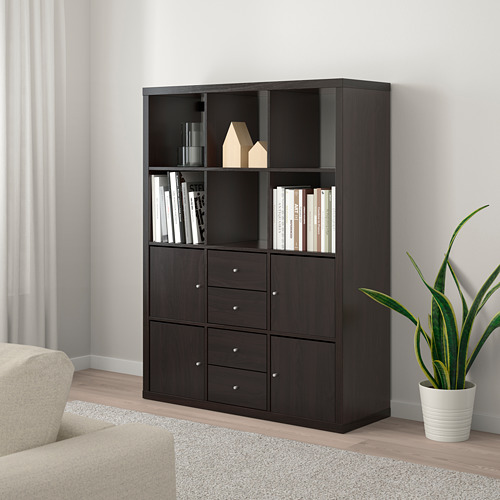 Estantería KALLAX negra con cajones y estantes, que almacena libros, jarrones y decoración junto a un sofá y una planta beige.