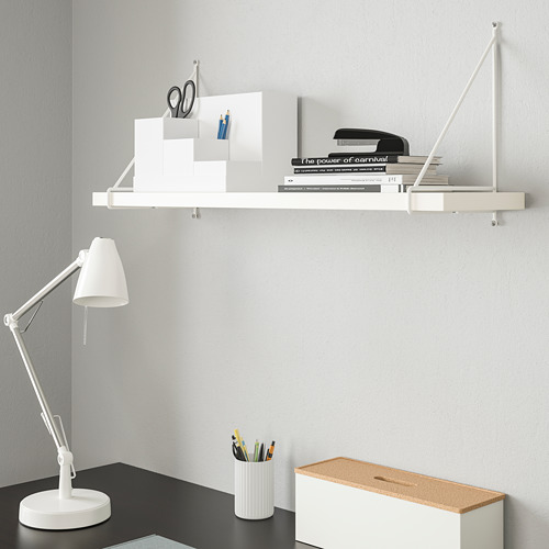 Estantería de pared blanca BERGSHULT con artículos de papelería, escritorio negro debajo con lámpara, taza para lápices y caja con tapa de corcho.