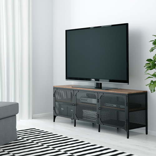Mueble de tv FJÄLLBO negro con tapa de madera, puertas de malla, pies ajustables, sosteniendo un televisor grande contra una pared blanca, con alfombra y sofá cerca.