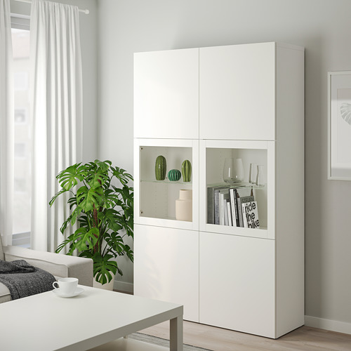 Gran armario blanco BESTÅ con puertas de cristal, para guardar objetos decorativos. Un diseño moderno y minimalista, colocado junto a una gran planta.