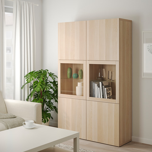 Armario de almacenaje de roble claro Ikea BESTÅ con puertas de cristal y estantes mixtos junto a mesa de centro y sofá.