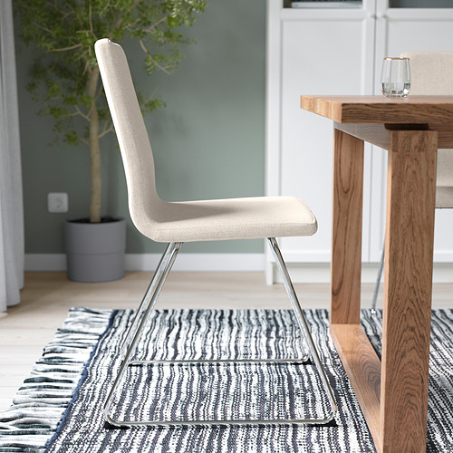 Silla moderna con tela beige, asiento y respaldo acolchados, base de patín cromada, sobre una alfombra de rayas blancas y negras cerca de una mesa de madera.
