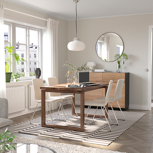 Moderno juego de comedor con mesa de madera, sillas beige, alfombra a rayas, aparador, espejo y lámpara colgante. Las ventanas proporcionan luz natural.