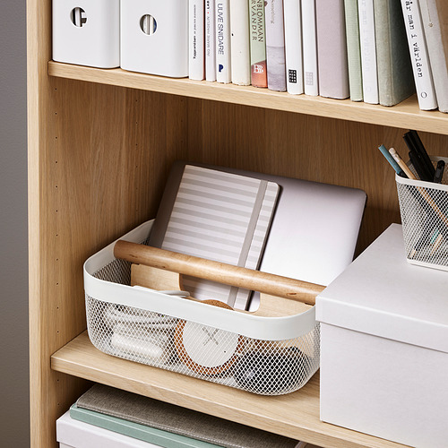 Estantería de madera con cesta de almacenamiento RISATORP que sostiene un ordenador portátil, un cuaderno y bolígrafos, ideal para organizar y transportar objetos pequeños.