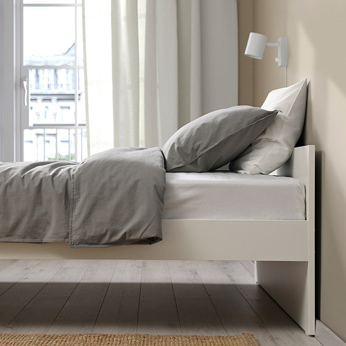 Un somier minimalista con ropa de cama gris y una lámpara de pared. Líneas limpias, diseño versátil y opciones de almacenamiento debajo.