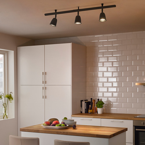 Cocina moderna con focos negros que iluminan la isla blanca repleta de frutas, platos y un jarrón. Gabinetes blancos y salpicadero de azulejos.