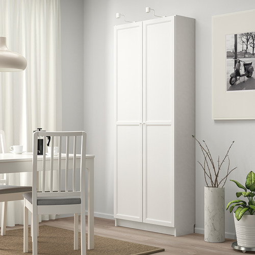 Una moderna estantería blanca BILLY se alza alta, cerca de una mesa de comedor y una silla, que ofrece un amplio espacio de almacenamiento. Su diseño elegante encaja bien en la luminosa habitación.