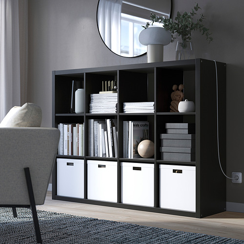 Sala de estar moderna con estantería KALLAX, libros, decoración y cajas. Acogedor rincón de lectura junto a sillón, espejo y lámpara.