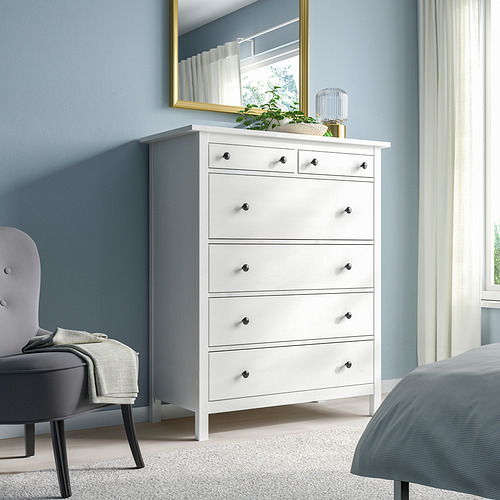 Cómoda blanca HEMNES de 6 cajones en pino macizo, estilo tradicional, alta y delgada, encaja bien junto a la cama y el espejo, con detalles dorados y verdes.