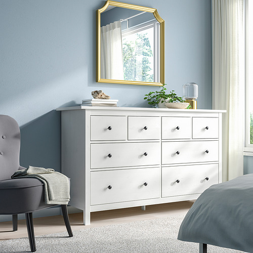 Dormitorio tranquilo con cofre HEMNES blanco, espejo grande, cama gris y silla. La cómoda de madera de seis cajones domina el espacio.