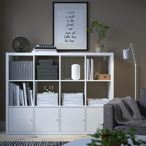 Estantería KALLAX con libros, papeles, decoración, póster, armarios cerrados. Alto, moderno, al lado del sofá y la lámpara.