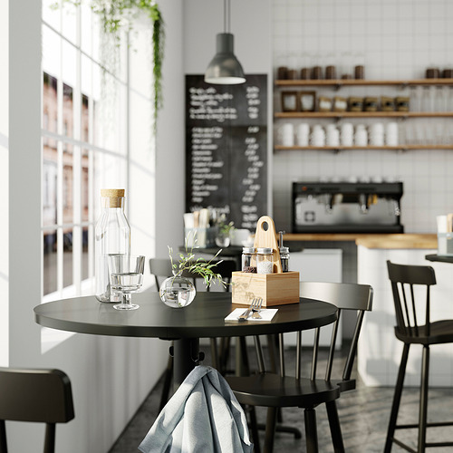 Cafetería moderna con mesa negra, sillas grises, organizador de condimentos de pino, cristalería, menú de pizarra y fondo blanco.