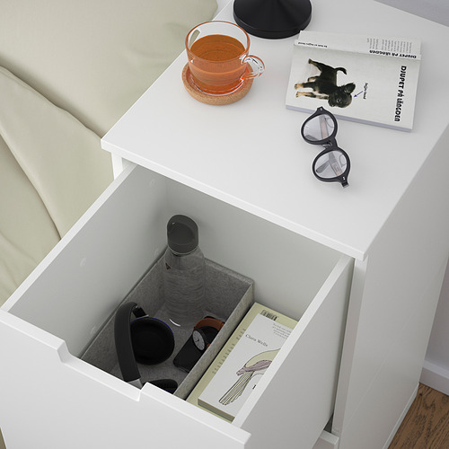 Cajón blanco de la mesita de noche abierto, mostrando una botella de agua, un reloj y libros. La parte superior tiene té, libro y vasos.