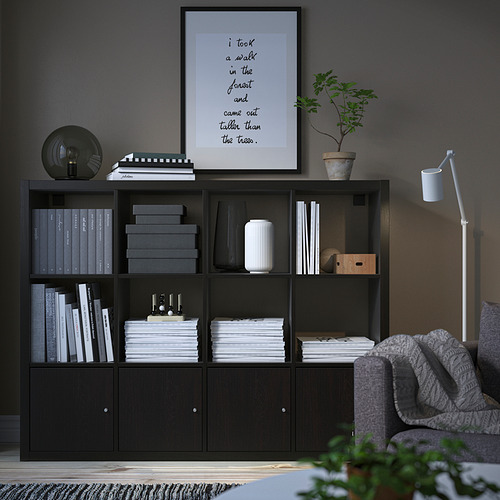 Estantería KALLAX negra con estantes y armarios, que contienen libros, marco de fotos, planta y decoración. Junto a un sillón gris.