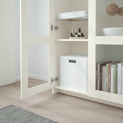 Mueble de almacenamiento BRIMNES blanco con puertas de vidrio, exhibiendo libros y decoración en estantes, y una caja blanca en el medio.