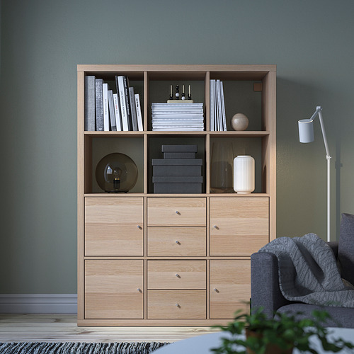 La estantería de roble claro KALLAX con cajones, que alberga libros, artículos decorativos y una lámpara, encaja bien en una acogedora habitación verde con un sofá gris cerca.