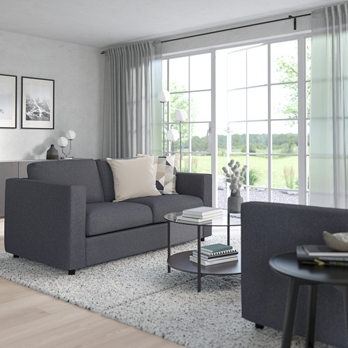 Sala de estar moderna con sofá gris VIMLE, cojines blancos, mesa de centro negra, gran ventanal y suelo de madera.