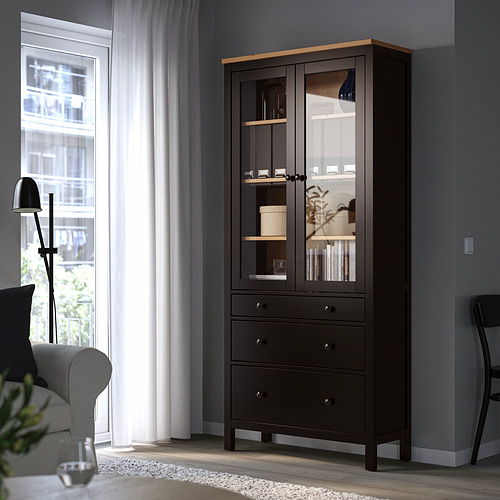La vitrina de madera negra HEMNES con puertas y cajones de vidrio se eleva junto a la ventana y la silla.