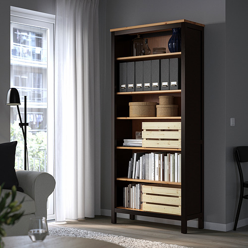 La estantería HEMNES de color marrón oscuro se alza en la esquina, llena de libros y contenedores de almacenamiento, junto a un sofá gris y una lámpara.
