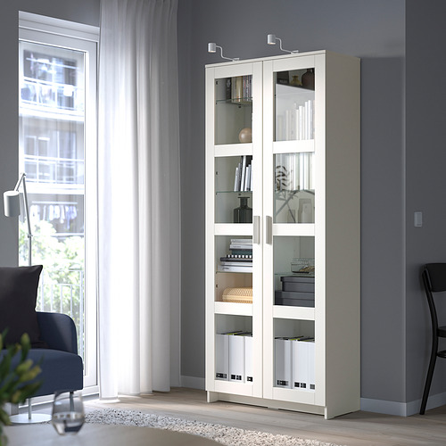 Mueble BRIMNES con puerta de cristal y estantes ajustables, cerca de grandes ventanales.