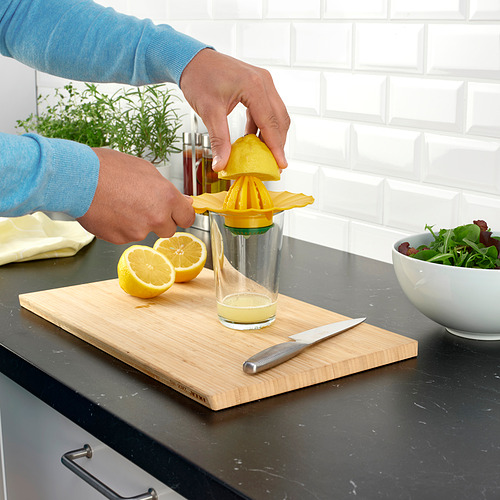 Persona exprimiendo limón con un exprimidor amarillo brillante en un vaso transparente, con mitades de limón cortadas y un cuchillo en una tabla de cortar de madera.