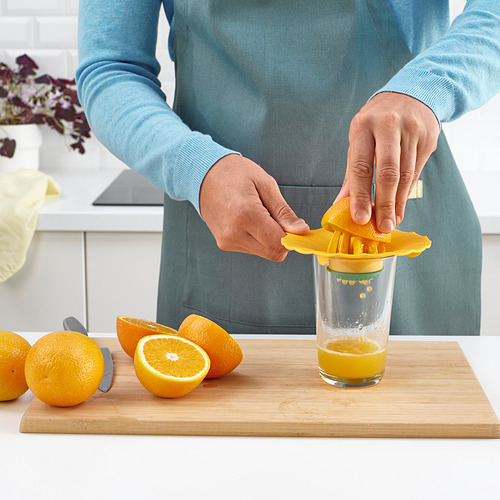 Persona exprimiendo jugo de naranja con un exprimidor de limón amarillo en un vaso sobre una tabla de cortar de madera. Naranjas enteras y cortadas por la mitad cerca.