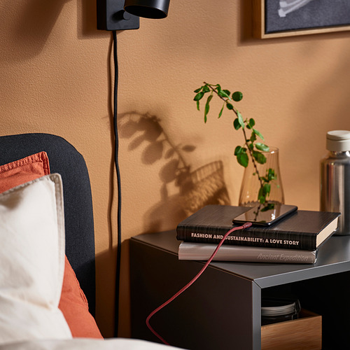 Configuración de cabecera con un cable USB LILLHULT negro que carga un teléfono encima de libros, cerca de una planta y una lámpara de pared.
