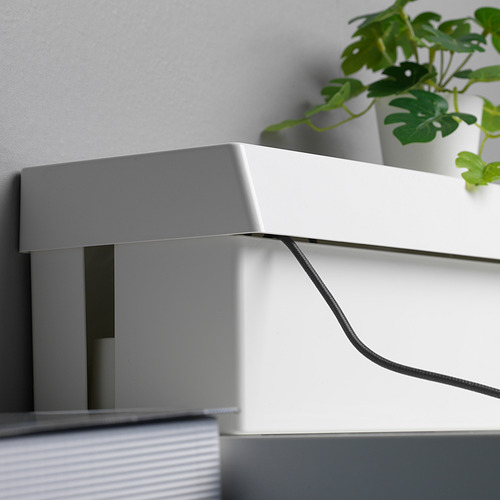 Caja blanca, probablemente el SÄTTING, cables ocultos. Tapa diseñada para facilitar el acceso a los cables. Planta cerca, cable visible.
