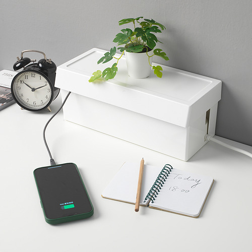 Un escritorio ordenado con una caja blanca que esconde cables, un teléfono de carga, una computadora portátil, un lápiz, una planta y un reloj.