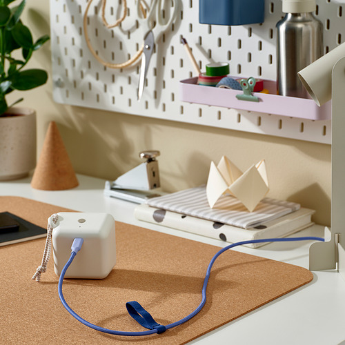 Un cable de carga USB-C blanco con un cordón azul y una banda elástica adjunta para un almacenamiento organizado, colocado en un espacio de trabajo de tapete de corcho.