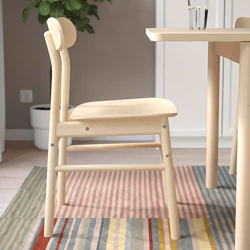 Silla de madera ligera con curvas suaves; se adapta a espacios clásicos y modernos; colocado sobre una alfombra a rayas junto a una mesa.