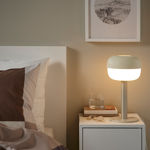 Junto a la cama con lámpara BLÅSVERK, almohadas blancas, vidrio y libros.