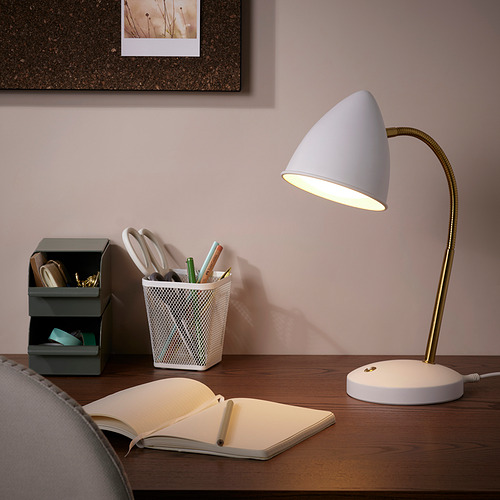 Lámpara ISNÅLEN en color blanco con cuello de latón sobre sobremesa. Cerca, un cuaderno abierto y un contenedor de papelería. Una luz suave ilumina el espacio de trabajo.