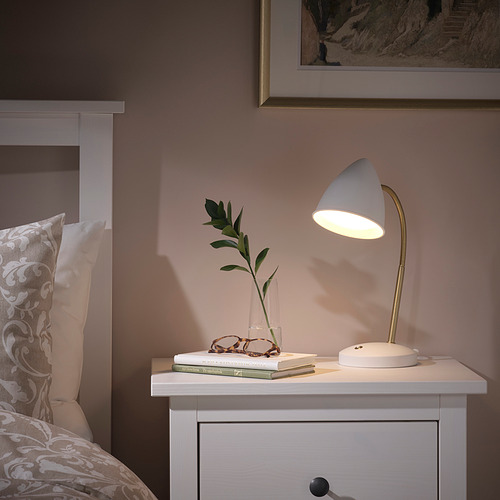 Lámpara de escritorio ISNÅLEN con cuello flexible junto a una cama, libros iluminados y un jarrón con plantas.