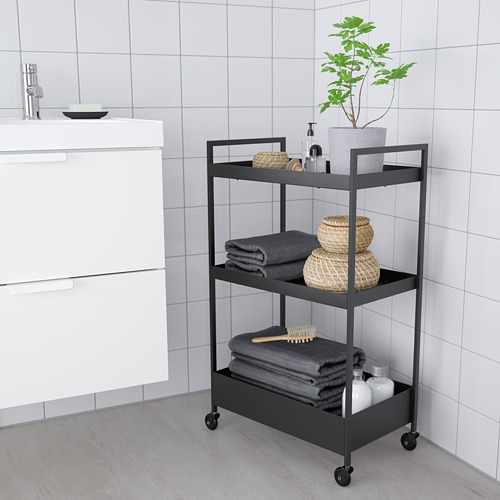 Carro nisaffors negro con 3 estantes, toallas de almacenamiento, cestas de mimbre y una planta. Cabe de forma compacta junto a armarios blancos.