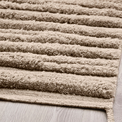 Primer plano de una alfombra de lana tejida a mano de felpa beige con patrón de ondas texturizadas sobre piso de madera.