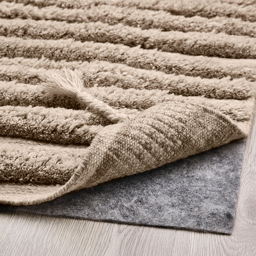 Alfombra peluda beige con patrón de ondas texturizadas, que muestra un pelo de lana grueso y una almohadilla antideslizante debajo.