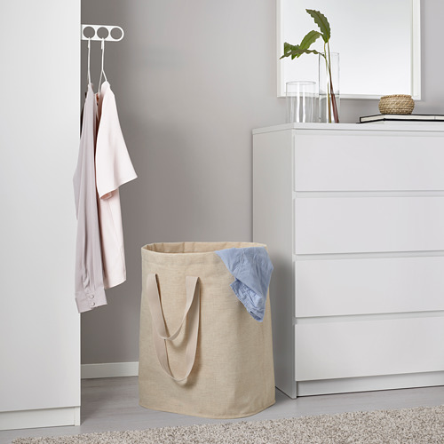 Bolsa de lavandería beige con asas cerca de la cómoda, sosteniendo la prenda azul sobre la alfombra.