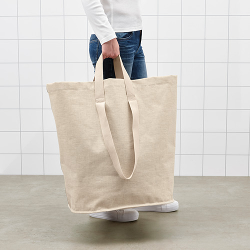 Persona sosteniendo una gran bolsa de lavandería beige con asas, de pie en una habitación con paredes blancas.