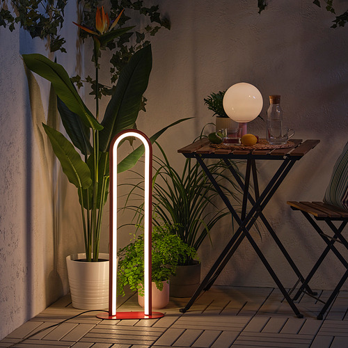 Moderna lámpara de pie iluminada en el patio exterior, que proporciona iluminación ambiental. Rodeado de plantas en macetas y una pequeña mesa con artículos.