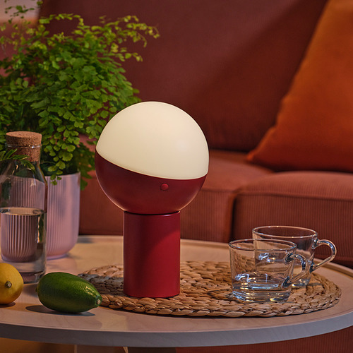 Lámpara de mesa moderna con pantalla de medio globo. Iluminando un espacio acogedor con una base roja. Rodeado de tazas y frutas en una mesa auxiliar.